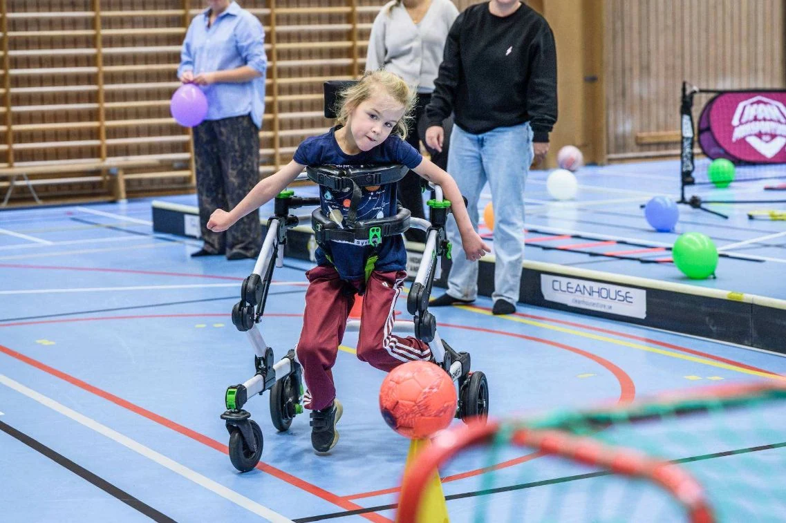 Ett barn med gånghjälpmedel spelar med en boll i en idrottshall tillsammans med ledare.